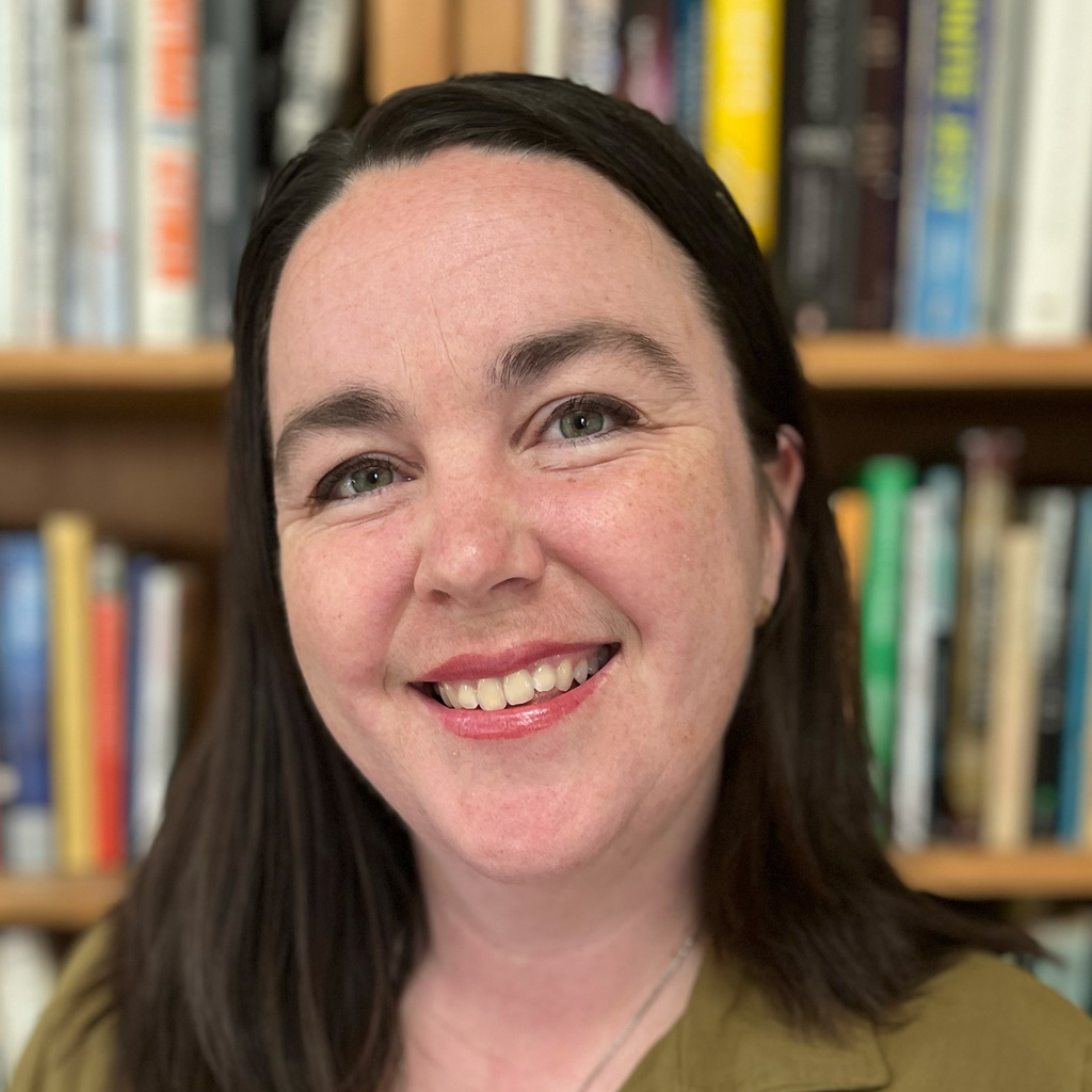 headshot of a woman with long brown hair standing in front of a bookshelf